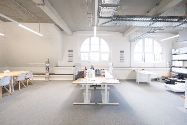 Ein modernes, lichtdurchflutetes Büro mit großen Fenstern, minimalistischen Möbeln und einer klaren, aufgeräumten Atmosphäre. Schreibtische und Stühle in hellen Farben sowie Pflanzen sorgen für eine angenehme Arbeitsumgebung.